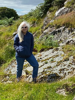 a woman in jeans and jacket poses midstride on a rugged hillside with water in the background