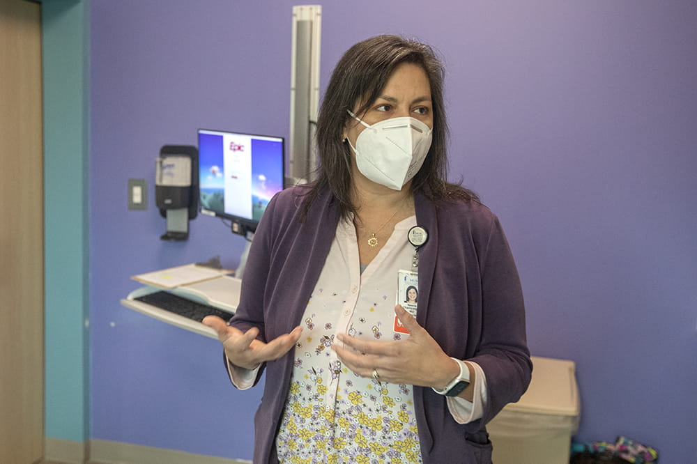 A woman with brown shoulder length hair wearing a surgical mask gestures as she talks. There is a computer screen behind her.