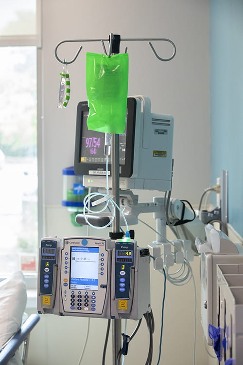 Machine in a clinic room. There is a monitor in back and a green bag filled with liquid hanging from a metal rack.