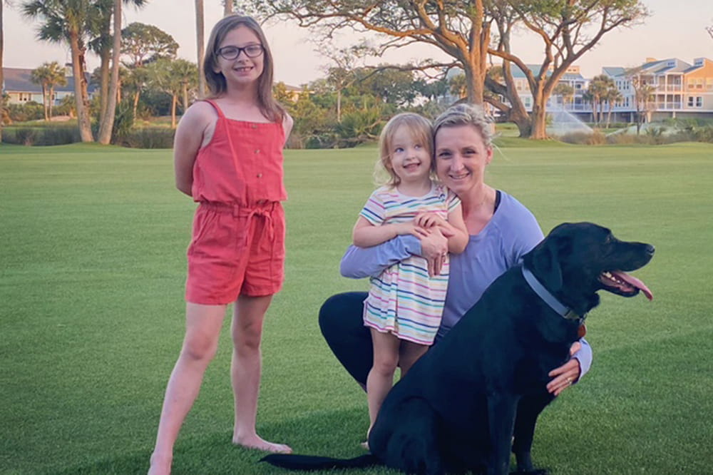 a woman kneels on a lawn holding a black Lab while a young girl leaning against her and an older girl standing nearby