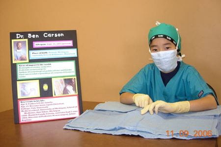 A young boy wearing scrubs, gloves and a mask sitting next to his poster on Dr. Ben Carson