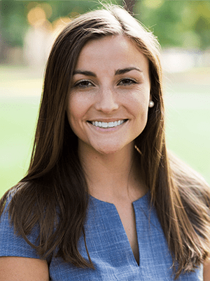 Headshot of young woman with long brown hair. She is smiling and wearing a blue short sleeved top.