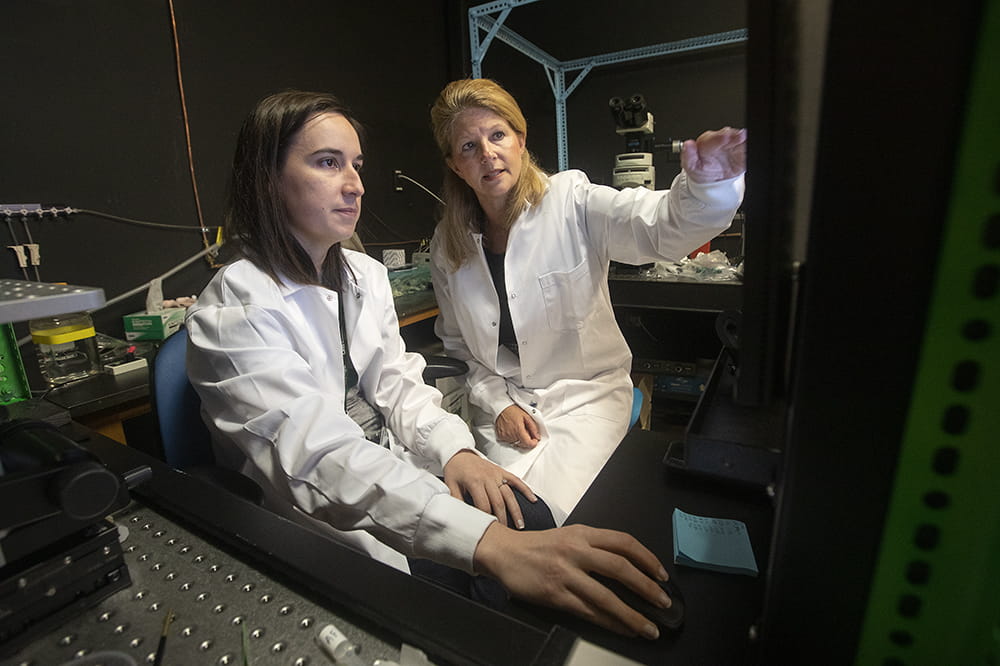 Two women in white lab coats sit close together. One is gesturing toward a screen. 