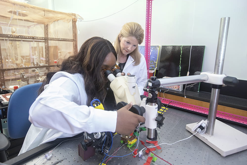 Two women in white lab coats use a microscope in a laboratory.