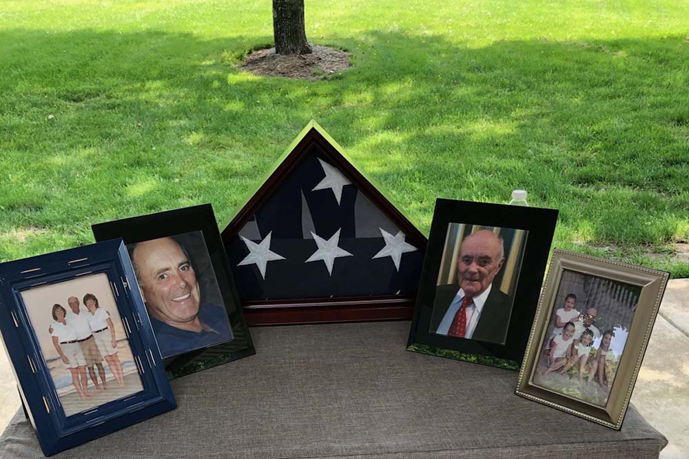 Four framed photos linked up on a table with a folded flag in the middle. They show a man and his family.