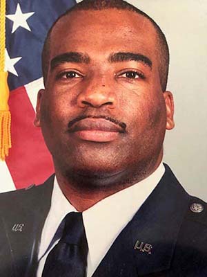 Headshot of a man in military uniform standing by an American flag. He has a mustache and closely cropped black hair.