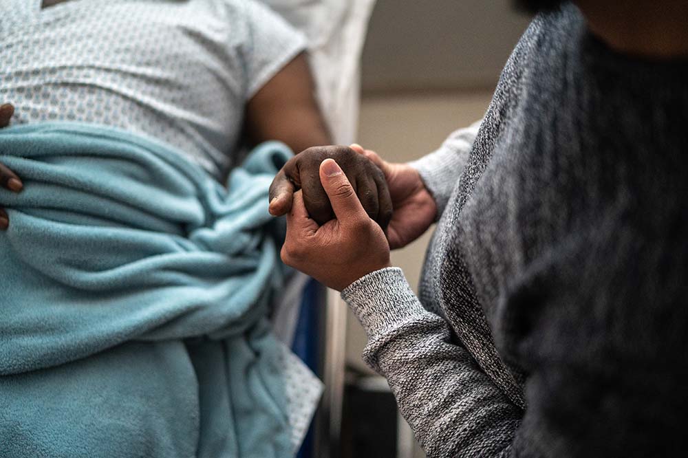 Closeup of two people's hands clasped. One is lying in a hospital bed. One is sitting beside the first.