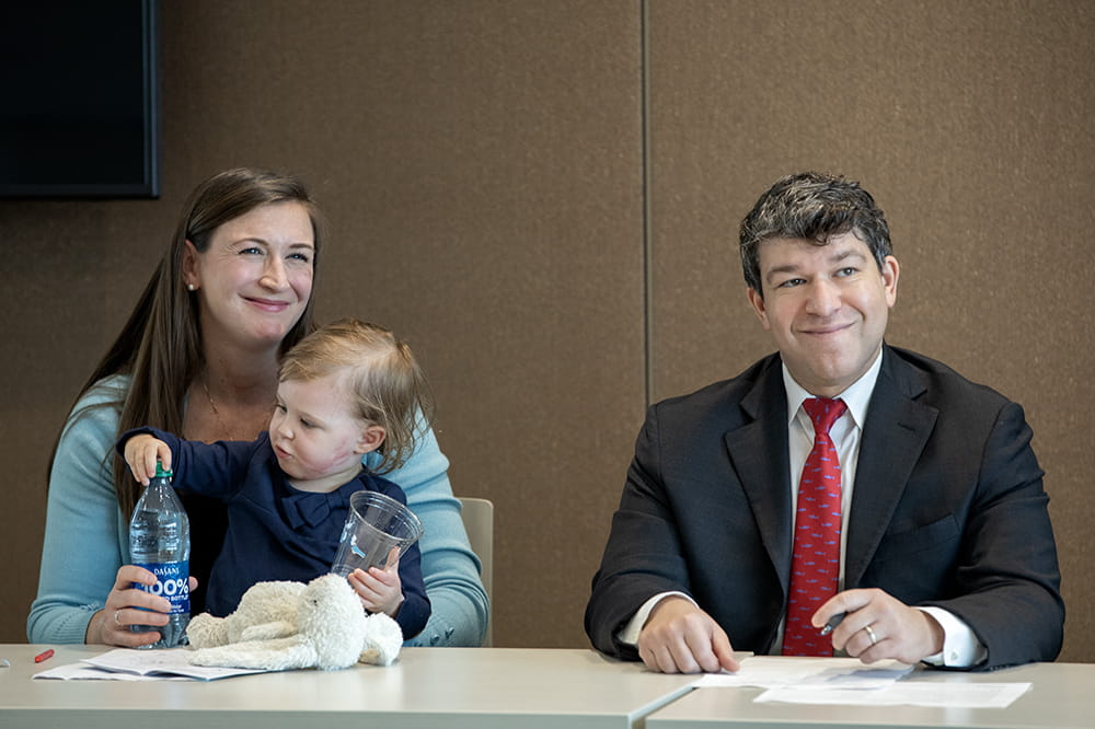 Kaitlin and Maeve Clark sit at a table with Dr. Steven Kahn. Kaitlin is smiling. Maeve is looking at a water bottle. Dr. Kahn is listening to a speaker.
