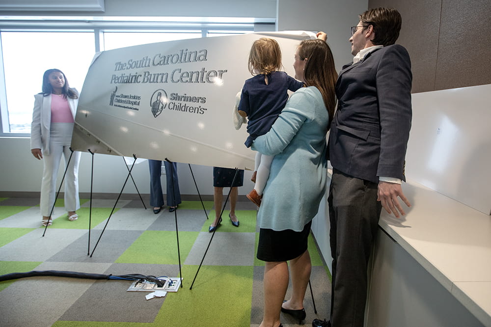 A young woman stands on the far left of a sign. A woman holding a girl and a young man are on the right.