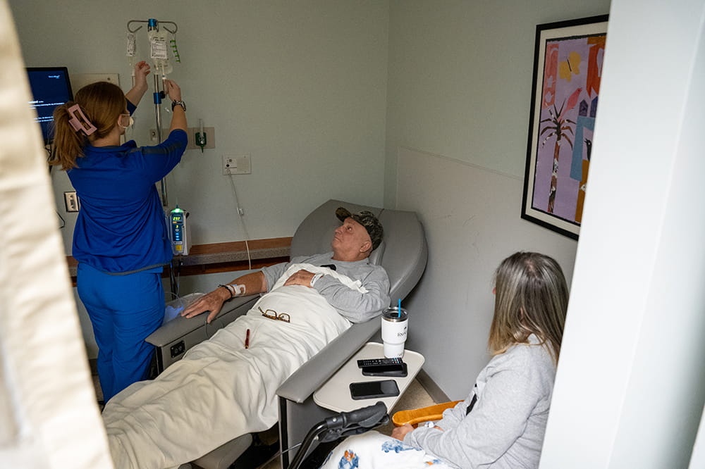 a nurse adjusts an IV line while a man in a reclining chair and a woman in a visitor's chair look on
