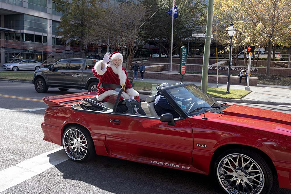 Santa waves while sitting on the back of a red convertible.