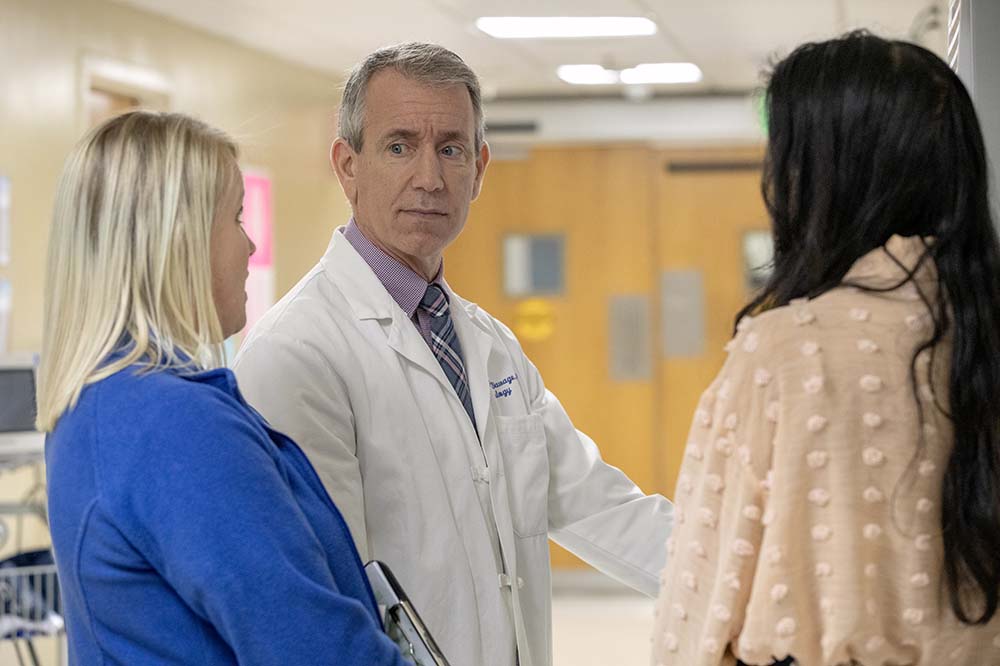 Doctor wearing a necktie and a white coat talks with two women in non-medical clothing in a hallway.
