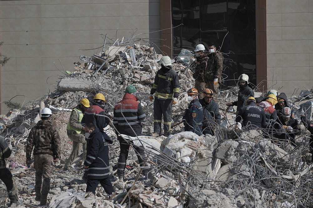 People in rescue uniforms stand on a dusty looking pile of rubble.