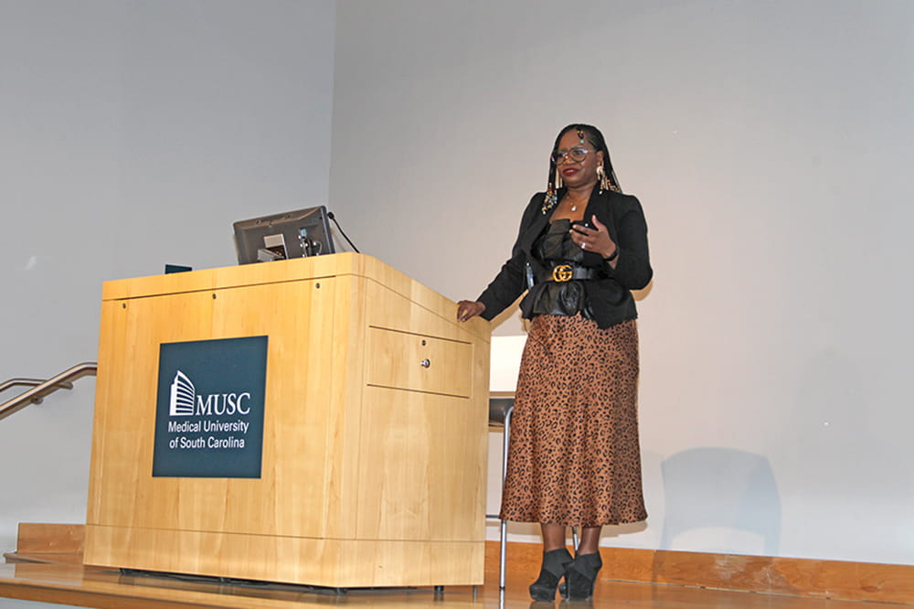 Woman speaks beside a podium. She is gesturing as she speaks about Black History Month. She is wearing a skirt and jacket.