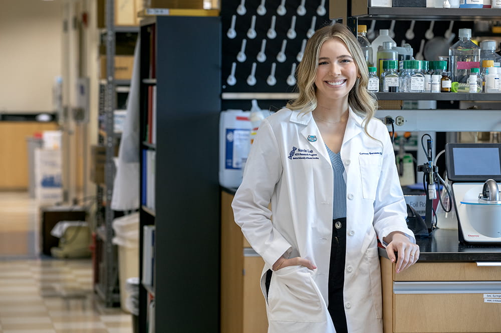 A woman with long blonde hair is standing in a lab wearing a white coat and
is smiling. 