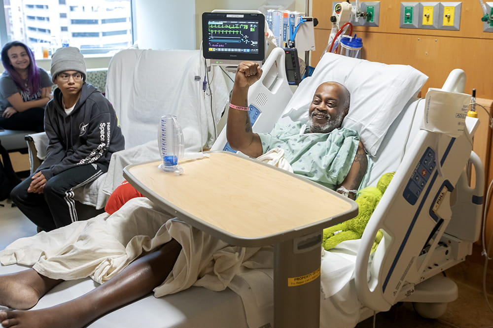 A man in a hospital bed gives a fist pump to someone out of frame