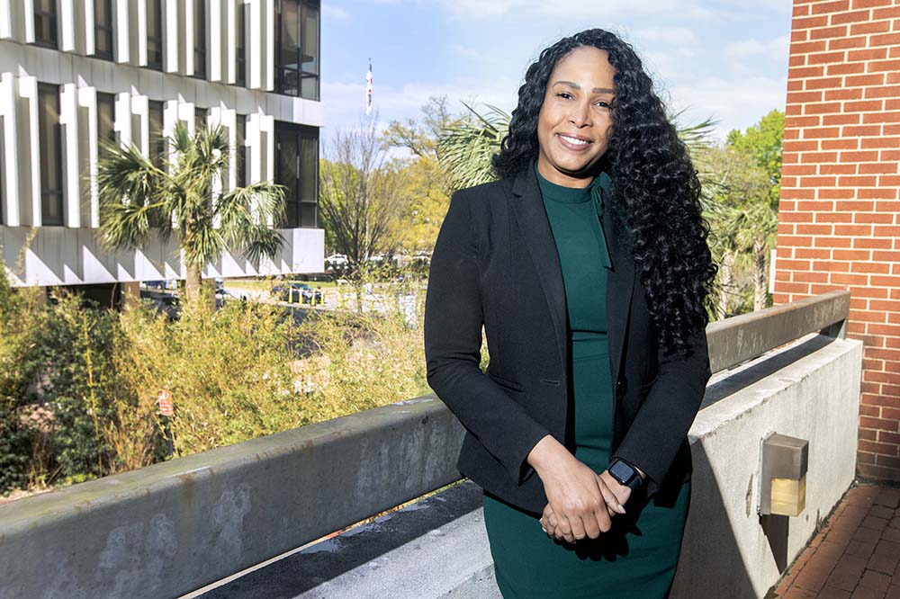 A woman with long, wavy black hair smiles as she stands on a porch outside. She's wearing a blazer and a green dress.