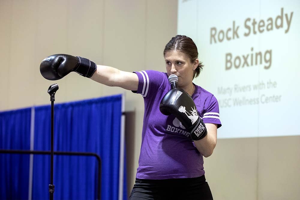 Woman with hair pulled back punches the air while wearing boxing gloves. A sign behind her says Rock Steady Boxing.