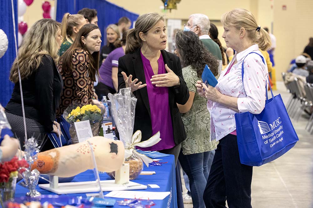 A woman wearing a white shirt with a blue MUSC bag over her shoulder talks with experts at the College of Nursing table about research.