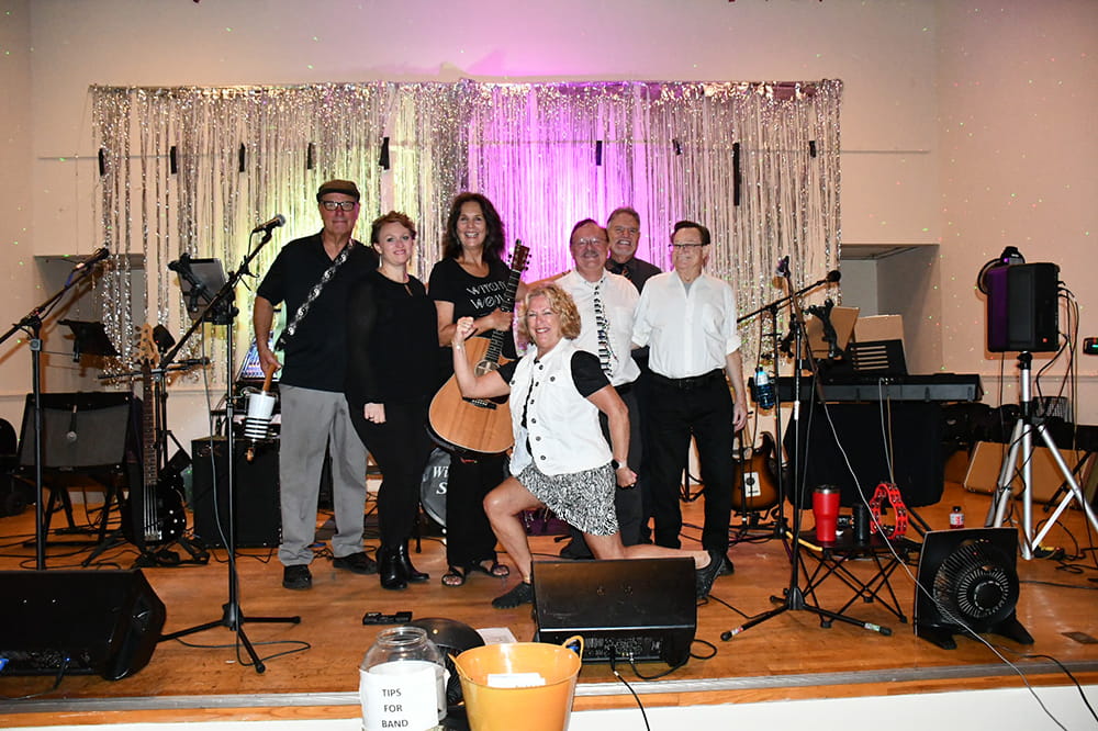 a woman jokingly makes a strongman pose in front of a group of bandmates on a stage filled with musical instruments