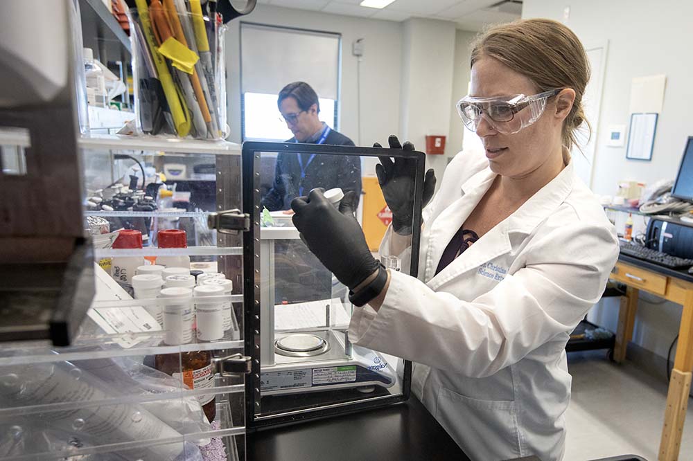 Woman wearing lab goggles and a white coat looks closely at the label of a vial.