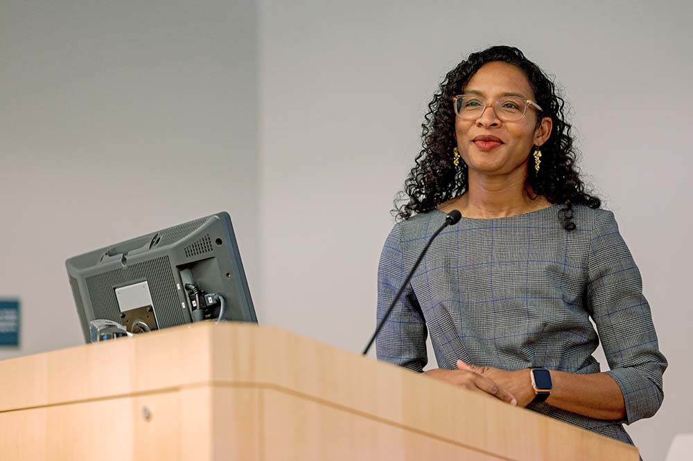 A woman wearing glasses with long, curly hair stands at a podium.