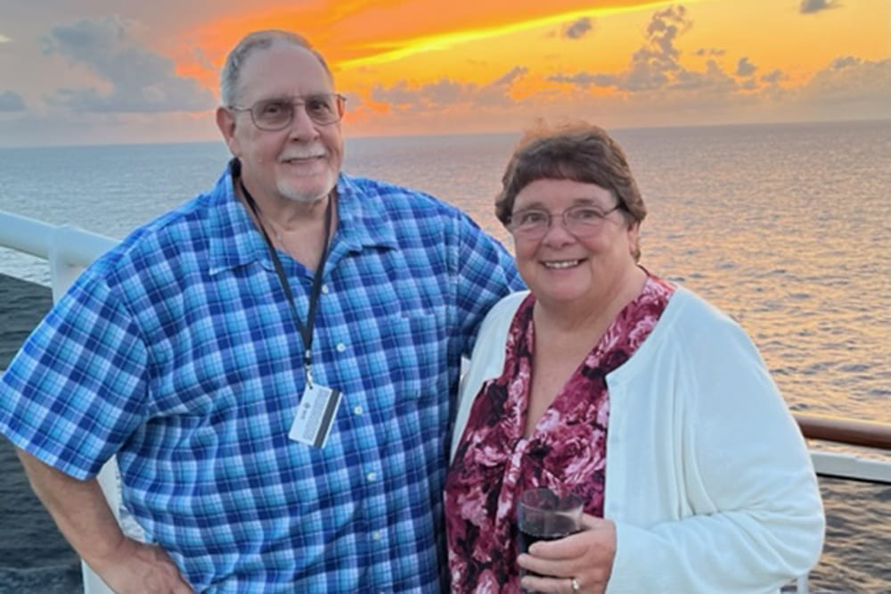 a couple poses on a cruise ship deck in front of a sunset