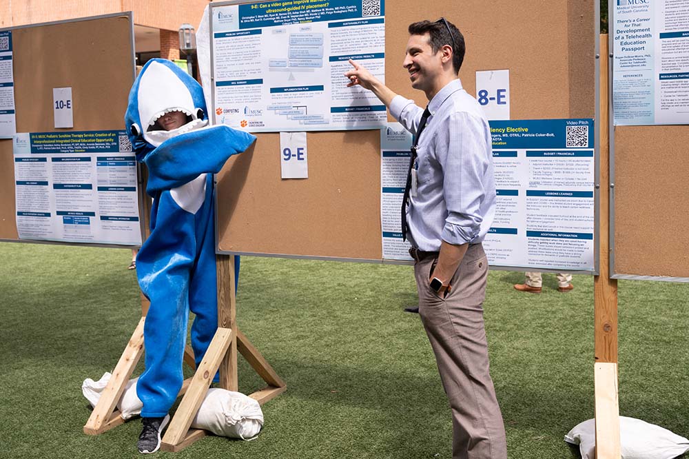 A person in a shark costume stands by a cork board with posters on it as a man wearing a necktie gestures to one of the posters.