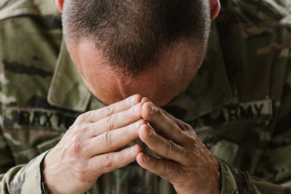 Man bowing head over steepled hands. He is wearing camouflage fatigues.