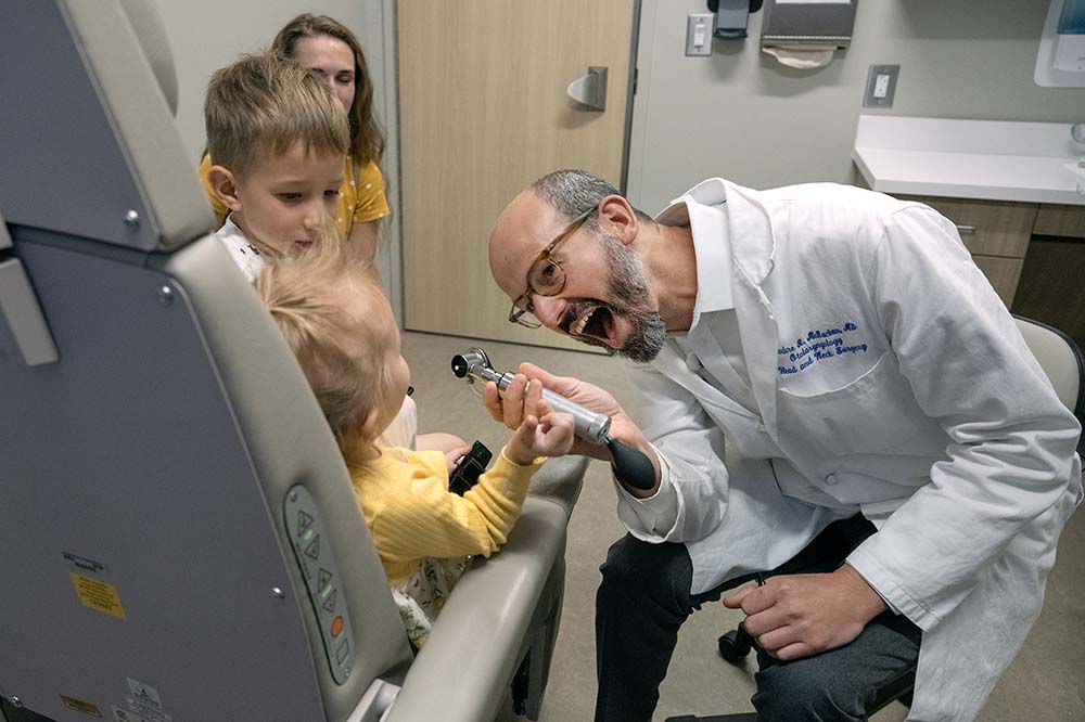 A smiling, bearded doctor holds a medical tool toward a little girl. You can see the back of her head. She is wearing a yellow top. A boy stands beside her watching.