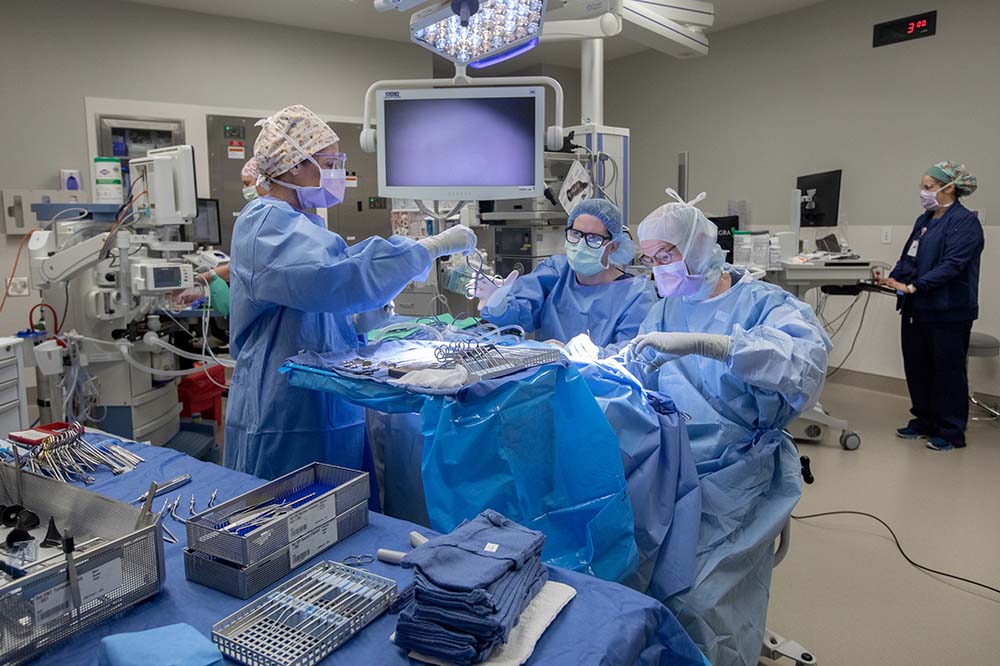 A team of people in blue scrubs and surgical masks works on a patient. A monitor hangs above them.