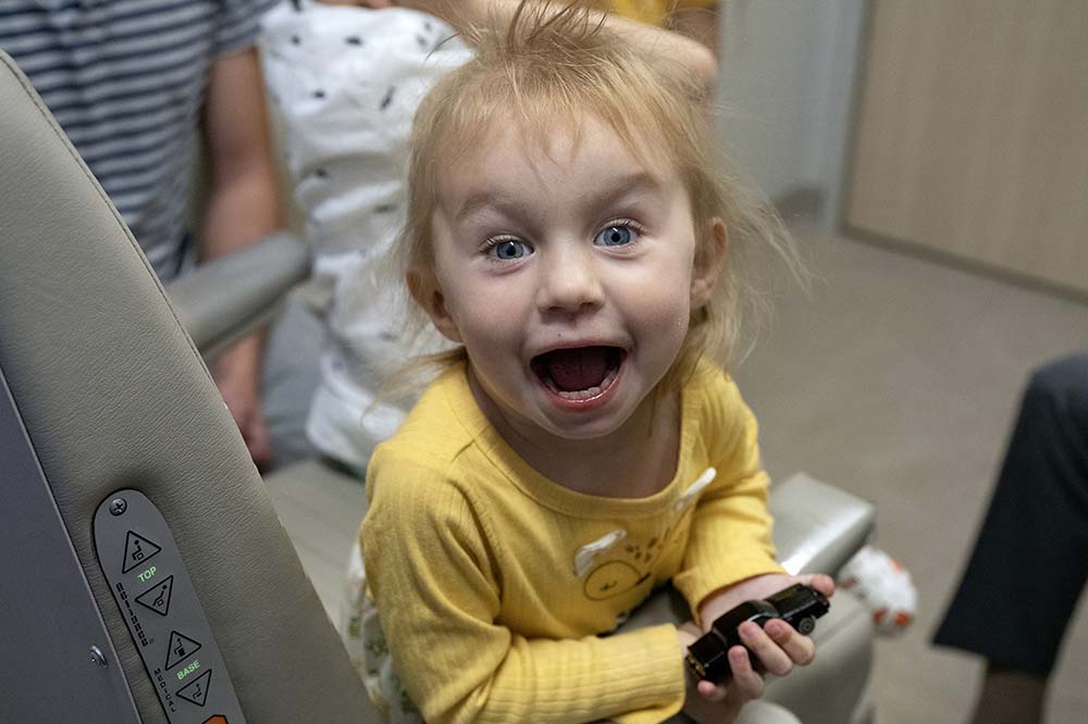 A little girl with blonde hair wearing a yellow top gives a surprised look with her mouth open.