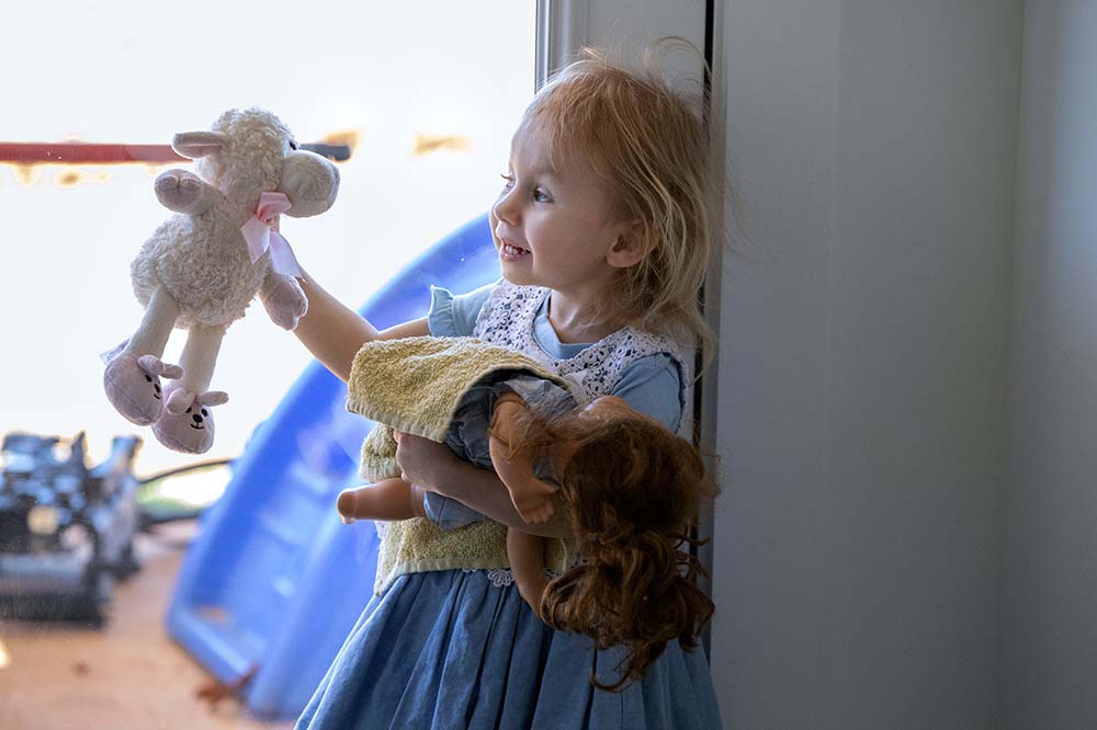 A little girl wearing a blue dress holds out a stuffed lamp. She's holding another toy as well.