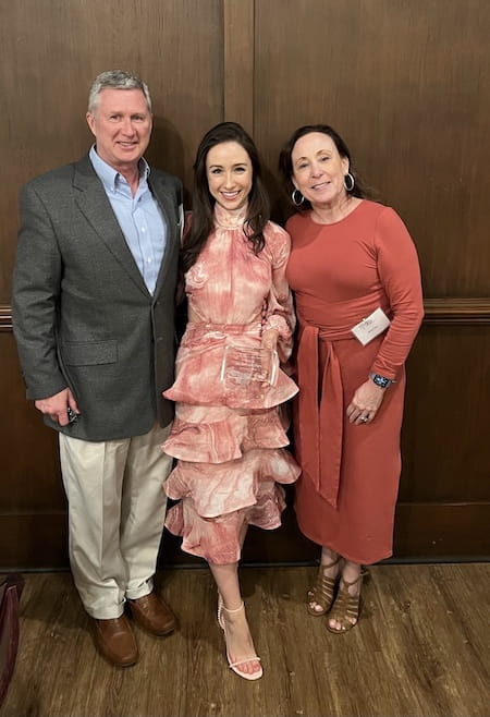 A man dressed in a suit jacket and khakis and woman in a dark pink dress bookend their daughter who is wearing a flowy pink and white dress