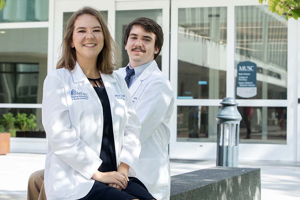 A sister and brother smile while wearing white coats in front of a building.