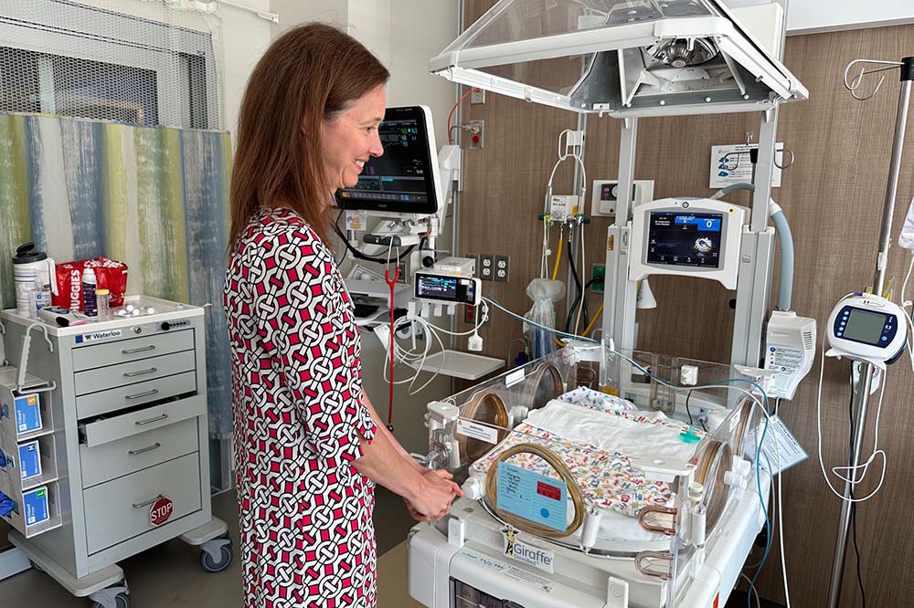 Woman in a print dress stands beside a baby's hospital bed. She is resting her hand on the edge of it.