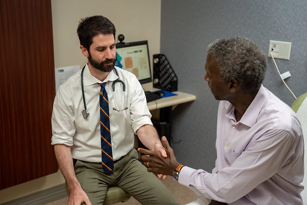 a doctor, sitting on a swivel stool, grasps a patient's left hand in his land hand