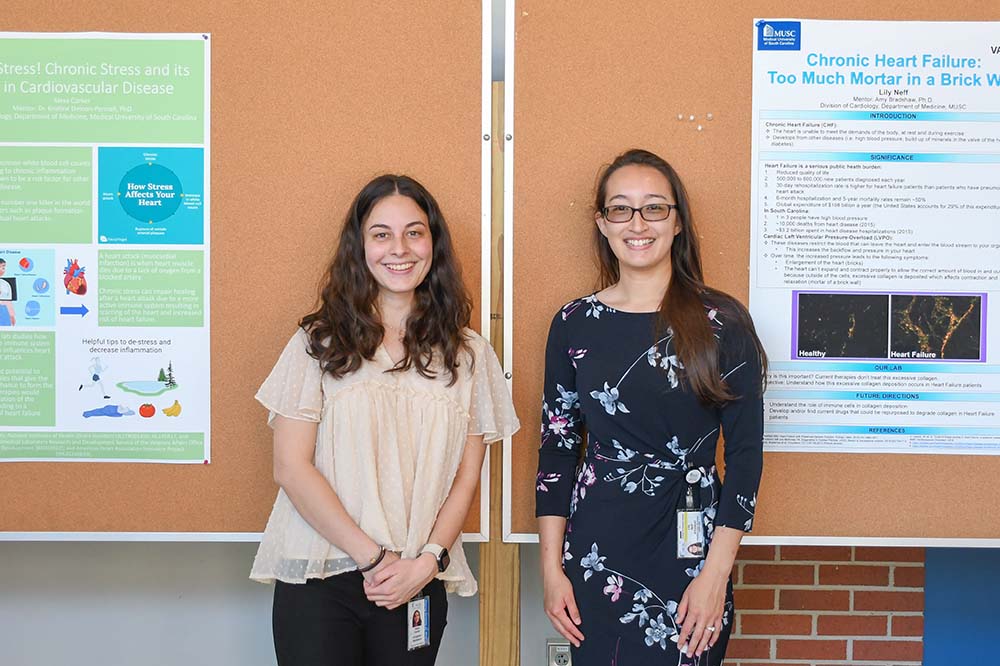 Two young women with long dark hair wearing business casual clothes stand in front of a cork board with two large posters on it.