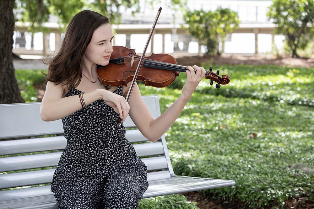Young woman with long brown hair is wearing a black jumpsuit and playing a violin on a bench outside.