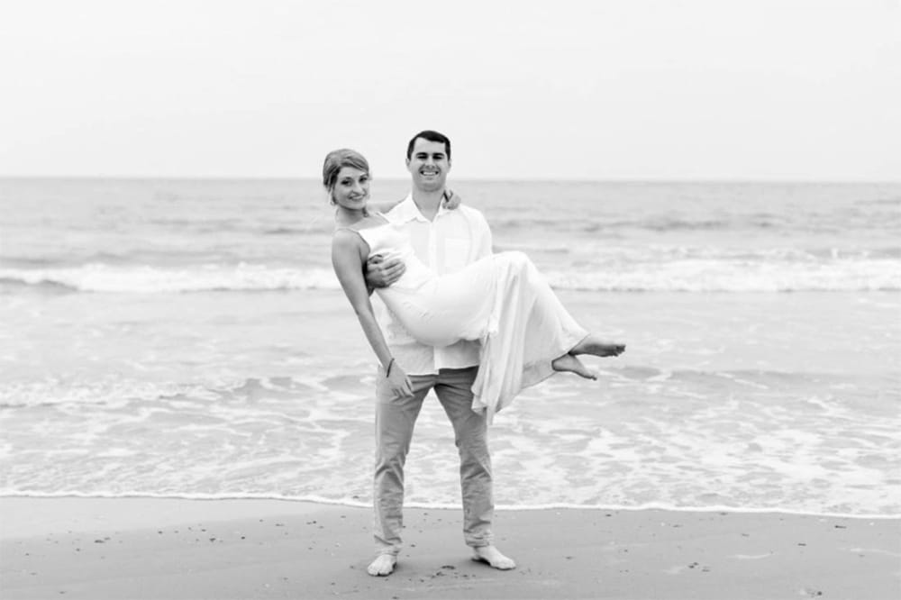 Zach holds his wife Katy in his arms while the couple stand on the beach. Zach is wearing a white shirt and khaki pants and Katy is in her wedding dress.