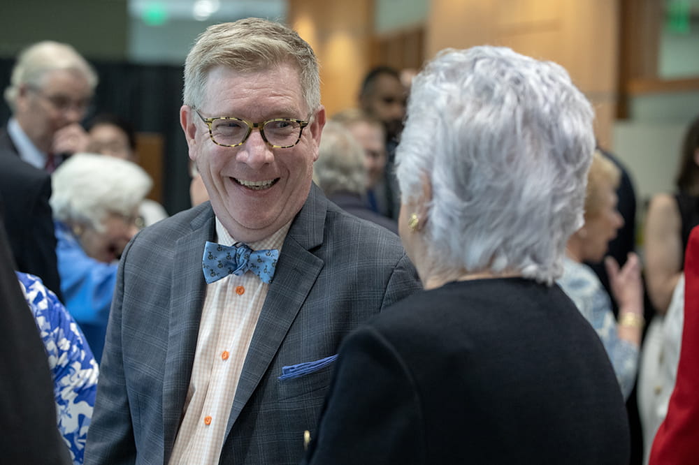 A man wearing round glasses and a bow tie laughs while facing a woman with white hair whose back is to the camera.
