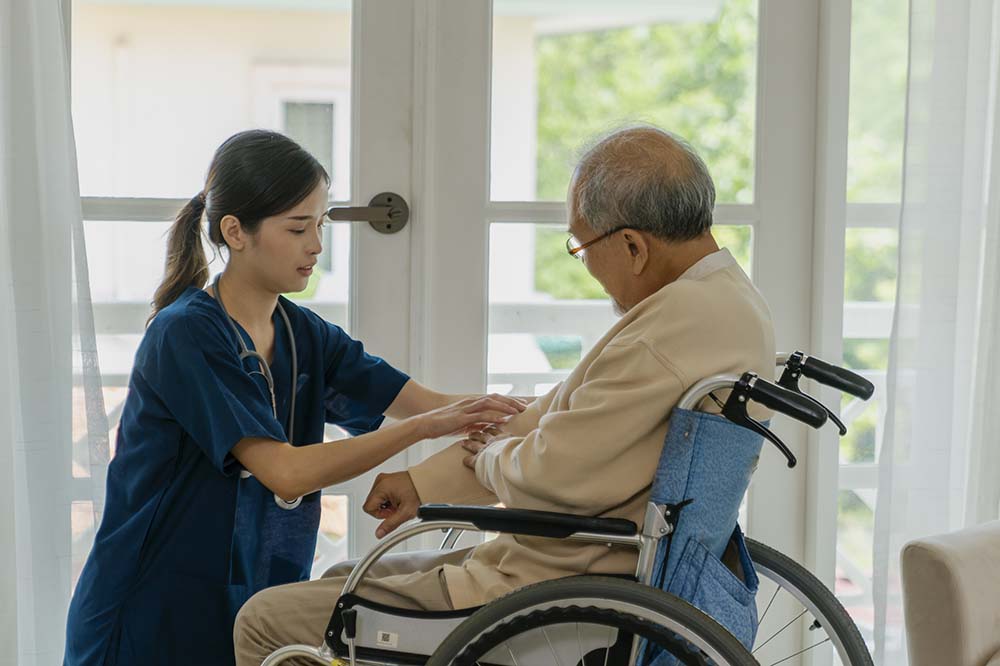 A woman in nurse's scrubs reaches out to a man in a wheelchair.