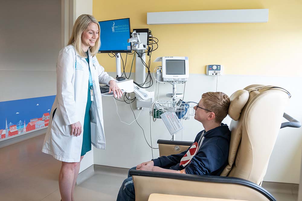 A blonde woman in a doctor's coat is standing in front of a seated boy wearing glasses.