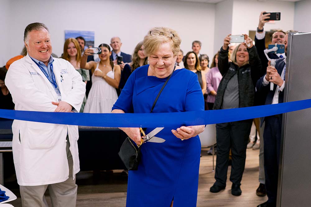 Smiling woman in a blue dress cuts a blue ribbon with a pair of scissors. People behind her are watching and smiling, too.