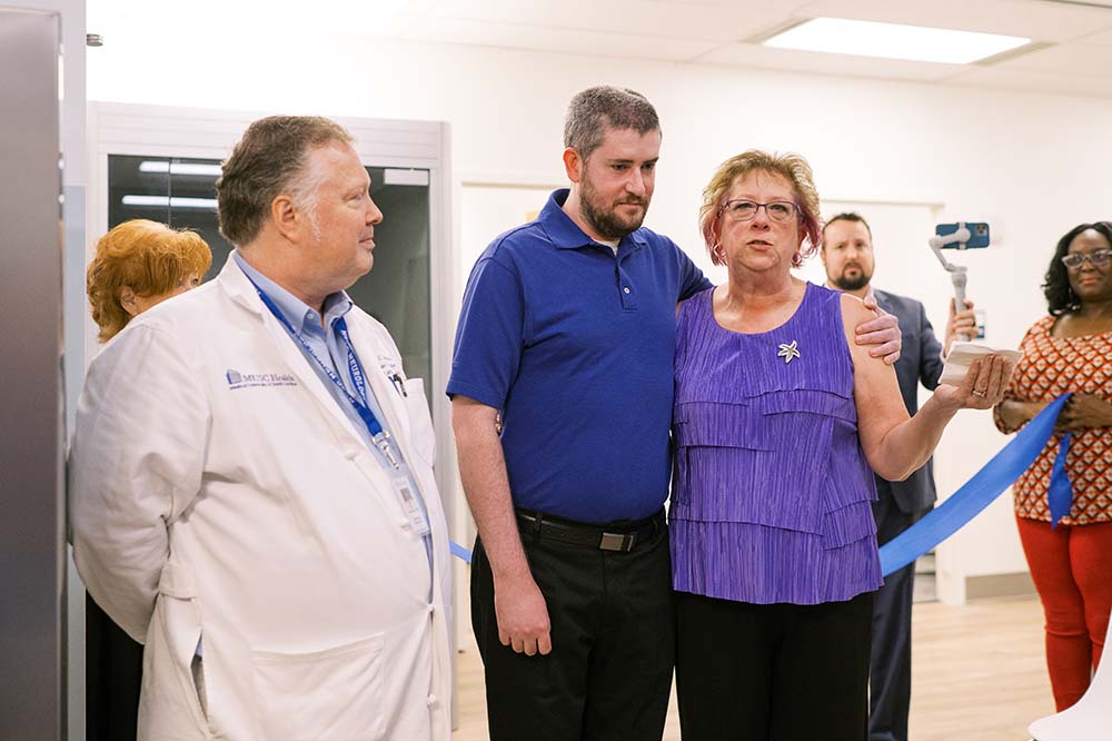 A doctor in a white coat listens as a woman wearing a purple top speaks. Her son is by her side. He has a beard and is wearing blue and black.