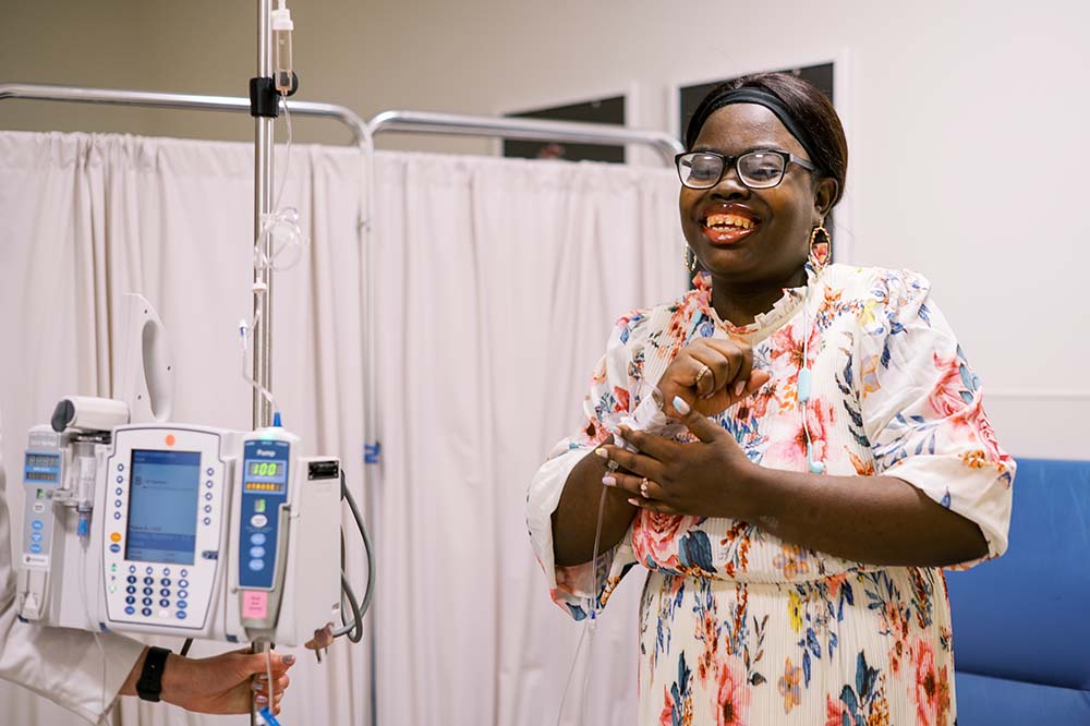 Woman wearing a floral dress and glasses smiles standing by an IV stand.