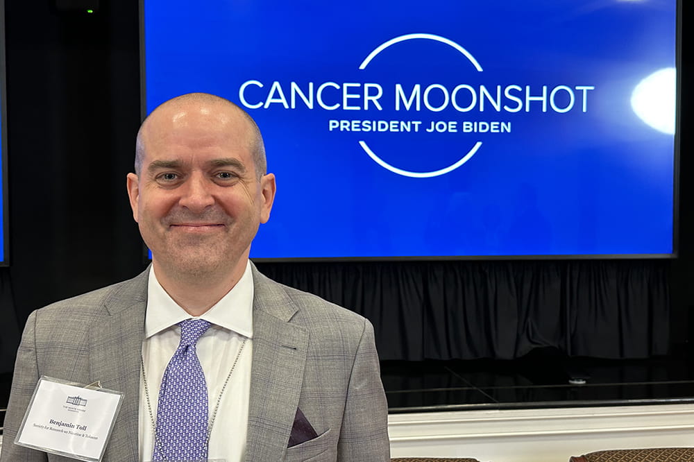 a man stands in front of a screen with the logo for the Cancer Moonshot