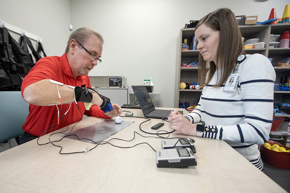 Man wearing a red shirt and glasses raises his gloved right hand. A woman sits across from with a computer screen in front her and wires connecting the man's glove with a monitoring system.