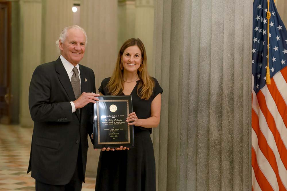 Man with white hair, wearing a suit and tie, holds a plaque with a woman. She has long, light brown hair and is wearing a short sleeved black dress. There is a United States flag beside them,