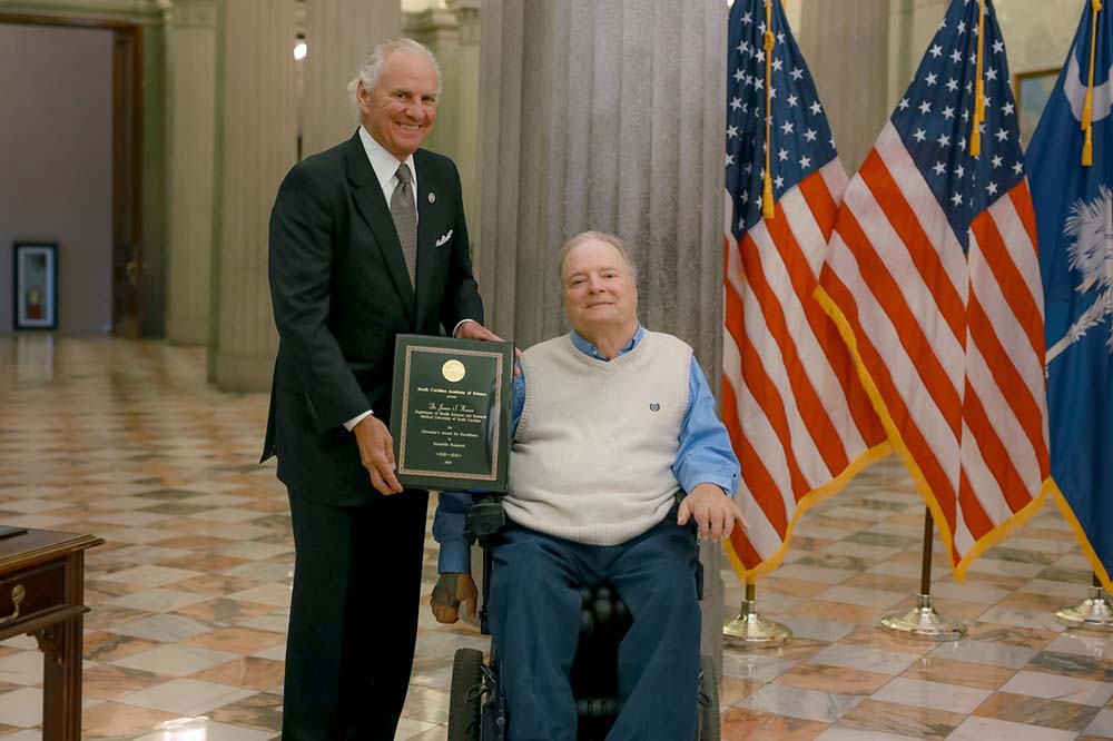 A man with white hair wearing a shit and tie stands beside another man who is in a wheelchair. The seated man is wearing a sweater vest, blue shirt and blue pants. They are holding a plaque and two American flags and one South Carolina flag are visible beside them.